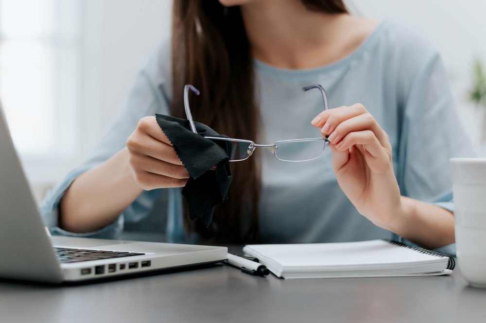 Nutzen Sie unterwegs am besten ein Brillenputztuch Frau putzt Brille am Laptop